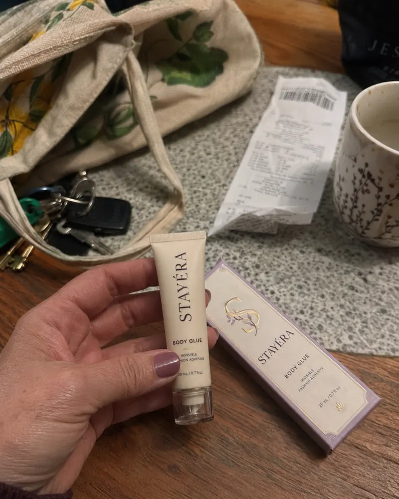 שפופרת STAYÉRA BODY GLUE ביד על שולחן מטבח לצד תיק בד, מפתחות ואריזת המוצר