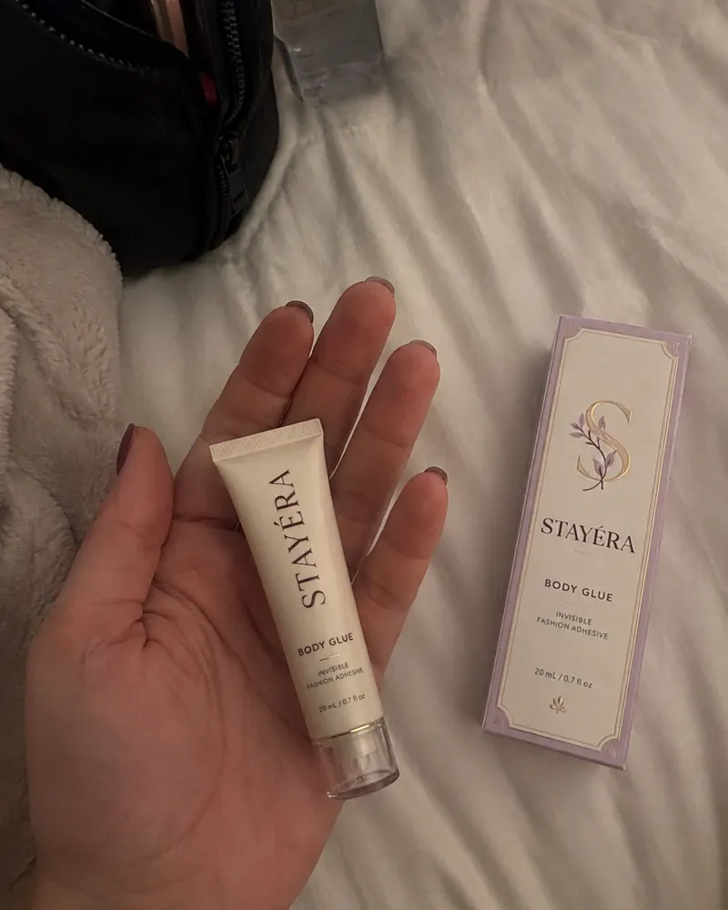 יד מחזיקה שפופרת STAYÉRA BODY GLUE לצד אריזת הקרטון על מיטה בהירה