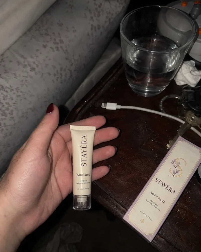 שפופרת STAYÉRA BODY GLUE ביד לצד שידת לילה עם כוס מים ואריזת המוצר